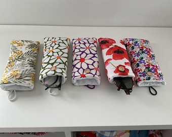May include: Five fabric eyeglass cases with various floral patterns. The cases are white with colorful designs, including yellow mimosa, green daisies, purple flowers, red poppies, and a multicolored floral print. Each case has a wooden button closure.