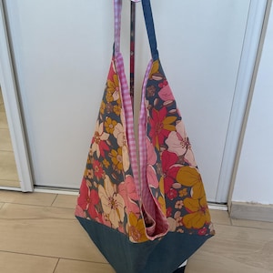 May include: A large tote bag featuring a floral pattern in pink, yellow, and peach on a dark grey background. The bottom of the bag is solid blue denim. The interior has a pink and white gingham pattern. The straps are blue denim. The bag hangs from a metal hanger.