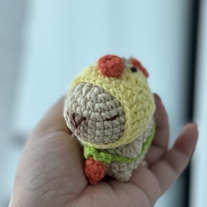 May include: A small crocheted stuffed animal wearing a yellow and orange hat and a green and orange outfit. The animal is brown and has a cute face.