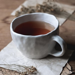 May include: A white ceramic mug filled with tea rests on aged paper. Dried flowers and paper with handwritten text surround the mug. The mug has a rounded shape and a handle. The tea appears to be a dark amber colour.