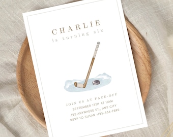 May include: A white invitation card with brown text announcing a birthday party for Charlie, who is turning six. The invitation features a watercolor illustration of a hockey stick and puck on a blue background. The text reads "Charlie is turning six. Join us at face-off September 15th at 11am 123 Anywhere St., Any City RSVP to Susan +123-456-7890"