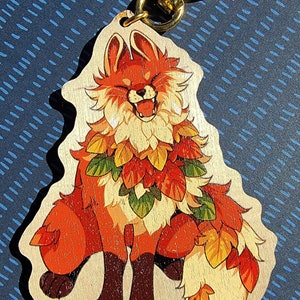 May include: A wooden keychain featuring a cartoon fox with bright orange and red fur, wearing a garland of autumn leaves in shades of orange, yellow, and green. The fox is smiling with its mouth open, showing its teeth. The keychain is attached to a gold metal ring.