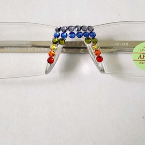 May include: Clear plastic frame glasses with rainbow colored rhinestones on the temples. The glasses have a silver metal bridge and the words "HARDENED LOCATED LENS APPROVED ABRASION RESISTANT TAIWAN" on a green sticker.
