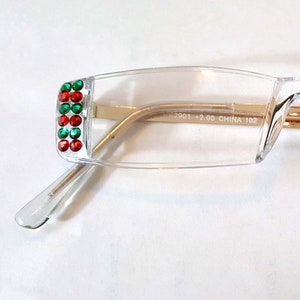 May include: Clear plastic framed glasses with red and green rhinestones on the temples. The glasses have a gold metal bridge and the words "Hardened Located Lens Approved Abrasion Resistant Taiwan" on a green sticker.