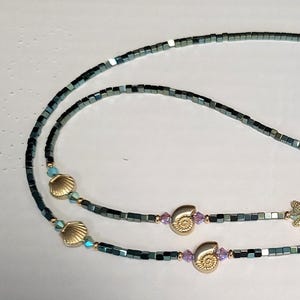 May include: A pair of glasses chain featuring a beaded design with seashell and starfish charms. The chain is made of dark gray beads and has gold-toned accents.