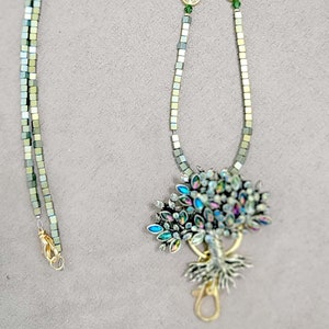 May include: A silver and green beaded lanyard with a gold tree of life charm. The charm has iridescent blue and green crystals.