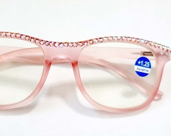 May include: Pink reading glasses with a clear lens and a rhinestone trim around the frame. The glasses have a blue sticker on the right side that says +1.25 Blue Light Filter.