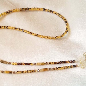 May include: A beaded eyeglass chain featuring a mix of amber and dark brown beads. The chain includes rectangular and small round beads, with silver-colored loops for attaching to glasses. The chain is designed to be worn around the neck.