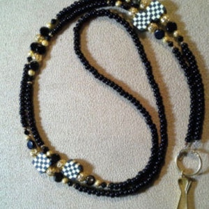 May include: Black and gold beaded lanyard with a checkerboard pattern charm. The lanyard has a metal clasp and a ring.
