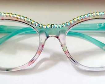 May include: Cat-eye reading glasses with a floral pattern and rhinestones around the frame. The lenses are clear and have a blue light filter. The glasses have a +2.00 magnification.