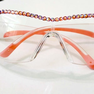 May include: Clear safety glasses with a pink ribbon design on the lenses. The glasses have a pink frame and a row of iridescent rhinestones around the top edge.