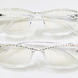 May include: Two pairs of clear plastic reading glasses with rhinestones on the frames. The glasses have a rectangular shape and the lenses are clear. The text +3.00 is printed on the right side of the frames.