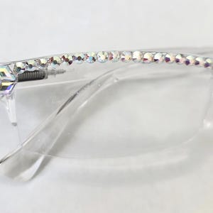 May include: Clear eyeglasses featuring a row of iridescent, round rhinestones along the top frame edge. A larger, square-shaped iridescent crystal is positioned at the temple hinge, adding a reflective detail to the transparent frame design.