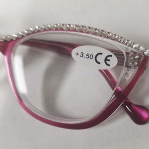 May include: Close-up of a pair of pink reading glasses with clear lenses. The top of the frames are embellished with small, clear rhinestones. A sticker on the lens indicates a +3.50 magnification. The glasses have a CE mark.