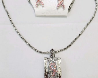 May include: A silver necklace and earring set. The necklace has a rectangular pendant with a textured frame and a pink ribbon design. The earrings are matching ribbon-shaped studs. The set is displayed on a white background.