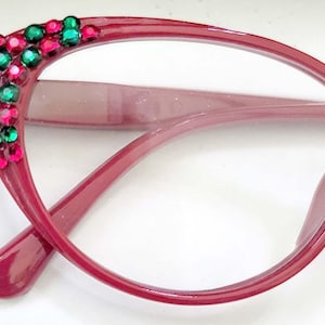 May include: Burgundy cat-eye glasses with clear lenses and a decorative cluster of green and pink rhinestones on the frame.