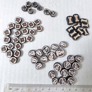 May include: A collection of polymer clay beads with dog-themed designs. The beads feature paw prints, dog faces, and bone shapes in brown, black, and cream colors. A ruler is visible at the bottom of the image.