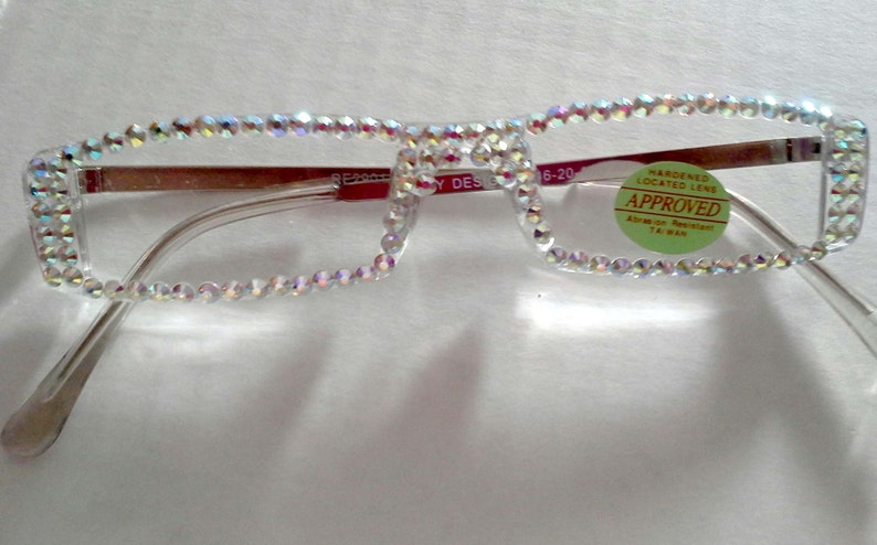 May include: Clear plastic framed glasses with a row of iridescent rhinestones around the edges of the frames. A green sticker with the text "HARDENED LENS LOCATED APPROVED" is on the right frame.