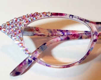 May include: A pair of cat-eye glasses with a floral print frame. The frame is a light purple and white with a pink and blue floral pattern. The glasses have clear lenses and are decorated with iridescent rhinestones on the top edge of the frame.
