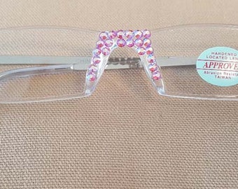 May include: Clear rectangular reading glasses with pink rhinestone accents on the temples. The glasses have a silver metal bridge and a sticker that says "HANDS-ON LOCATED LENS APPROVED ASIAN RESISTANT TAIWAN".