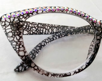 Può includere: Occhiali grigi da gatto con un motivo a pizzo nero e una fila di strass iridescenti attorno al bordo.