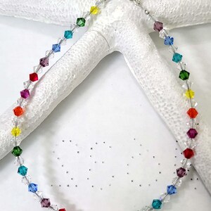 May include: A rainbow-colored beaded necklace with faceted crystal beads. The necklace features a spectrum of colors, including red, orange, yellow, green, blue, and purple. The necklace is displayed on a white, textured surface.