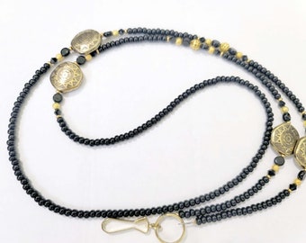 May include: A black beaded lanyard with gold accents and three gold metal charms. The charms have a sunburst design.