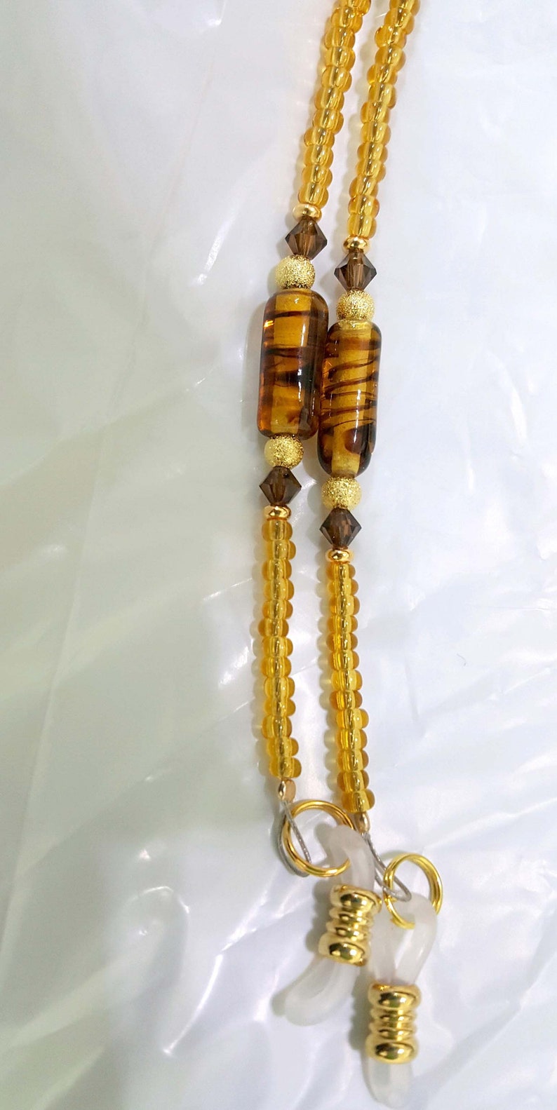 May include: A pair of gold-toned eyeglass chains with clear plastic ends and brown and gold beads. The chains are made of small, round beads.