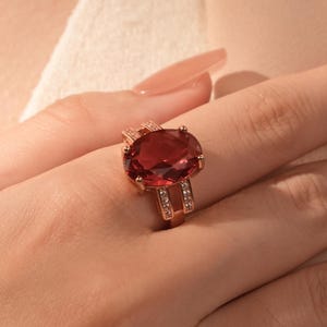 May include: A rose gold ring featuring a large, oval-cut, red gemstone. The ring has a detailed band with small, clear accent stones. The ring is displayed on a hand with long, polished nails, against a soft, neutral background.
