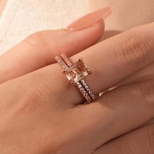 Puede incluir: Juego de anillos de compromiso de oro rosa con una piedra preciosa de color melocotón de corte cuadrado y una delicada banda de pequeñas piedras brillantes. La banda a juego también tiene pequeñas piedras.
