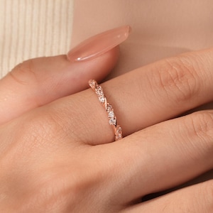 Peut inclure: Bague en or rose au design délicat. La bague présente de petites pierres précieuses transparentes disposées en motif répétitif. La bague est portée sur une main avec de longs ongles polis. L'esthétique générale est élégante et minimaliste.