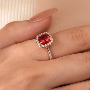 May include: A rose gold ring with a large, square-cut, red gemstone, encircled by smaller, clear stones. The ring's band is also set with small, clear stones. The ring is shown on a hand.