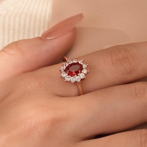 May include: A rose gold ring featuring a large oval red gemstone surrounded by smaller clear gemstones.