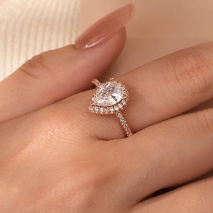 May include: A pear-shaped diamond engagement ring with a halo setting in rose gold. The ring is set with smaller diamonds around the centre stone.