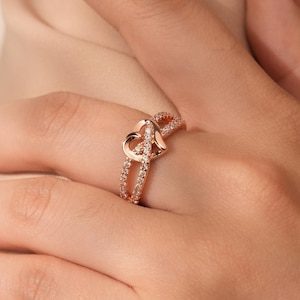 Può includere: Un anello in oro rosa con un design a doppio cuore. L'anello è incastonato con piccole pietre trasparenti.