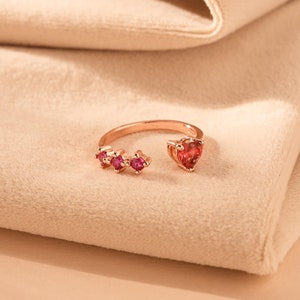 Può includere: Un anello in oro rosa con una pietra rossa a forma di cuore e pietre rosse più piccole sulla fascia.