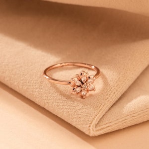 Peut inclure: Une bague en or rose avec un délicat motif floral serti de pierres brillantes transparentes. La bague est présentée sur un fond en tissu beige doux.