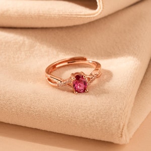 May include: A rose gold ring featuring a pink oval gemstone and smaller clear gemstones set in a twisted band.
