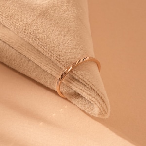 May include: A thin rose gold ring featuring a delicate twisted or diagonal textured pattern along its band. The metal has a polished finish that reflects light, creating subtle highlights across the surface of the circular jewellery piece.