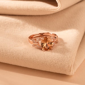 Può includere: Un anello in oro rosa con una grande pietra di topazio ovale circondata da diamanti più piccoli. L'anello è posto su un tessuto morbido color beige.