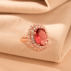 Può includere: Un anello in oro rosa con una grande pietra ovale rossa circondata da un doppio alone di piccole pietre chiare.
