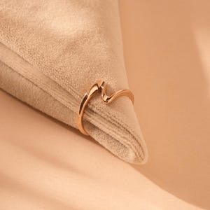 Puede incluir: Un anillo de oro rosa pulido con un diseño liso y continuo en forma de onda. La banda tiene un acabado metálico elegante y reflectante, con una curva única y suave que crea una silueta ondulada a lo largo de la parte superior del anillo.