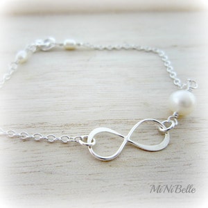 May include: A silver infinity charm bracelet with white pearls. The bracelet is on a white wooden surface.