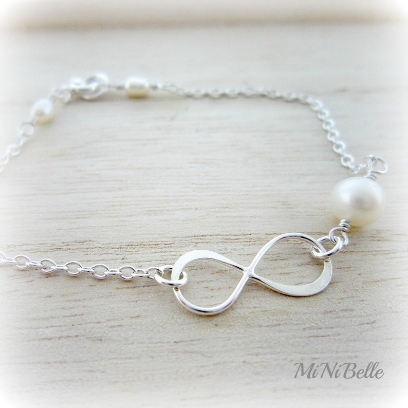 May include: A silver infinity charm bracelet with white pearls. The bracelet is on a white wooden surface.