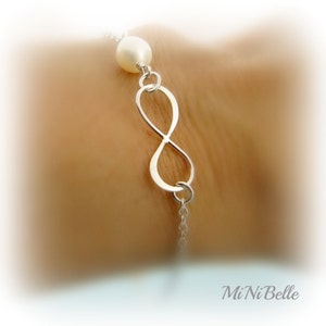 May include: A silver infinity symbol charm bracelet with a white pearl accent. The bracelet is on a person's wrist.