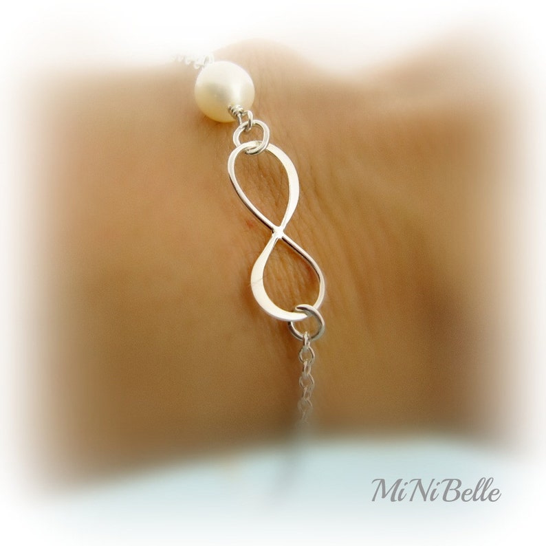 May include: A silver infinity symbol charm bracelet with a white pearl accent. The bracelet is on a person's wrist.