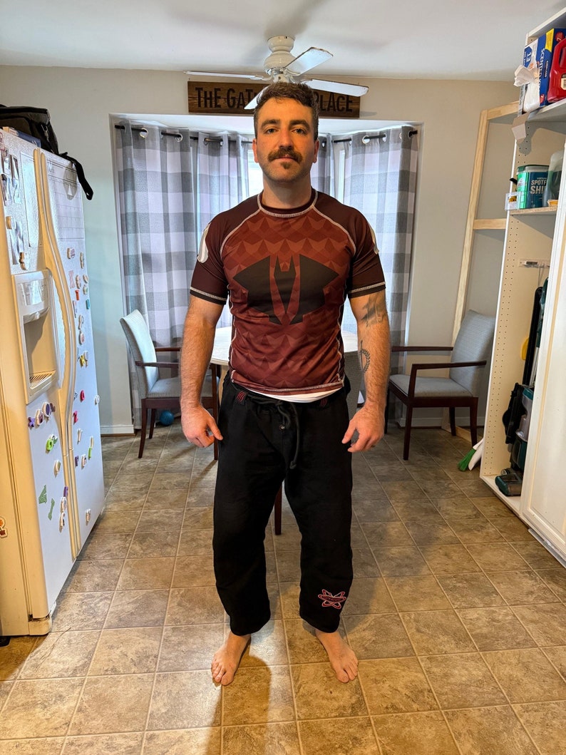May include: A man wearing a maroon rash guard with a black and white logo and black pants with a pink logo. The rash guard has a geometric pattern. The man is standing in a kitchen with a white refrigerator and a window behind him.
