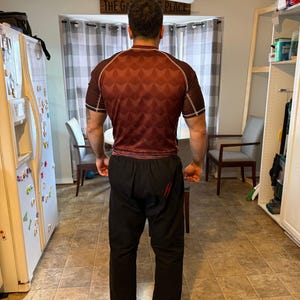 May include: A person wearing a red and black rash guard with a geometric pattern and black pants with a red stripe down the side. The text "THE GA" and "PLACE" is visible on a sign above the person.