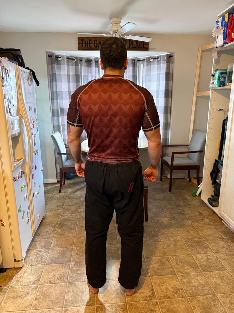 May include: A person wearing a red and black rash guard with a geometric pattern and black pants with a red stripe down the side. The text "THE GA" and "PLACE" is visible on a sign above the person.