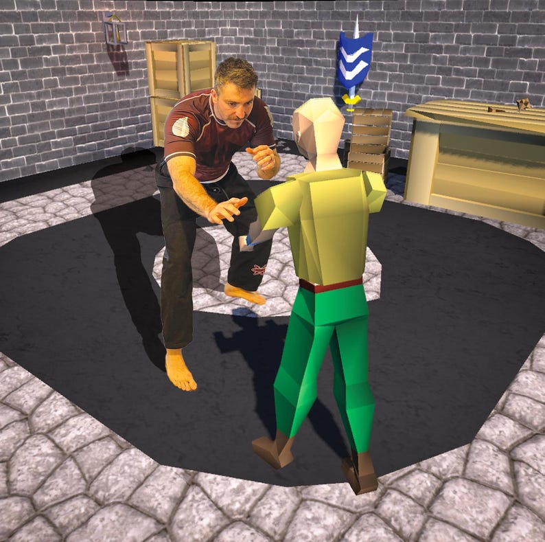 May include: A man in a red and black shirt is demonstrating a martial arts move to a green and yellow figure. The figure is standing on a black mat in a room with brick walls.